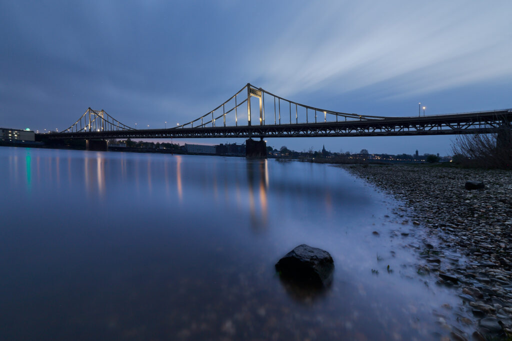 Beleuchtete Krefeld-Uerdinger Brücke, mit Spiegelungen im Rhein. Unscharf im Vordergrund liegt ein größerer Stein